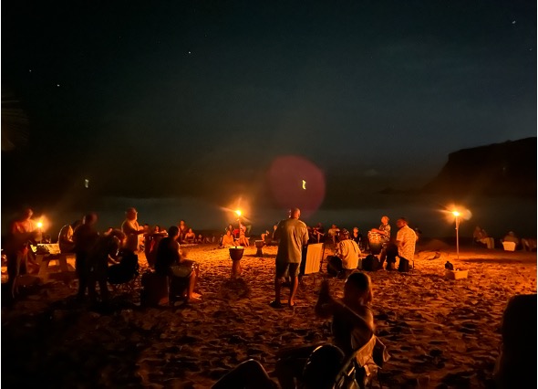 Círculo de participantes tocando tambores en la playa, iluminados por antorchas y rodeados de la vibrante energía de la música. La playa y las olas del mar se hacen visibles a lo lejos, creando una atmósfera de unión y serenidad.